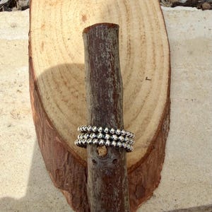 Puede incluir: Tres anillos plateados con cuentas apilados sobre un palo de madera marr&oacute;n oscuro, sobre un fondo de madera marr&oacute;n claro. Los anillos est&aacute;n hechos de peque&ntilde;as cuentas redondas. La madera tiene un aspecto natural y texturizado.