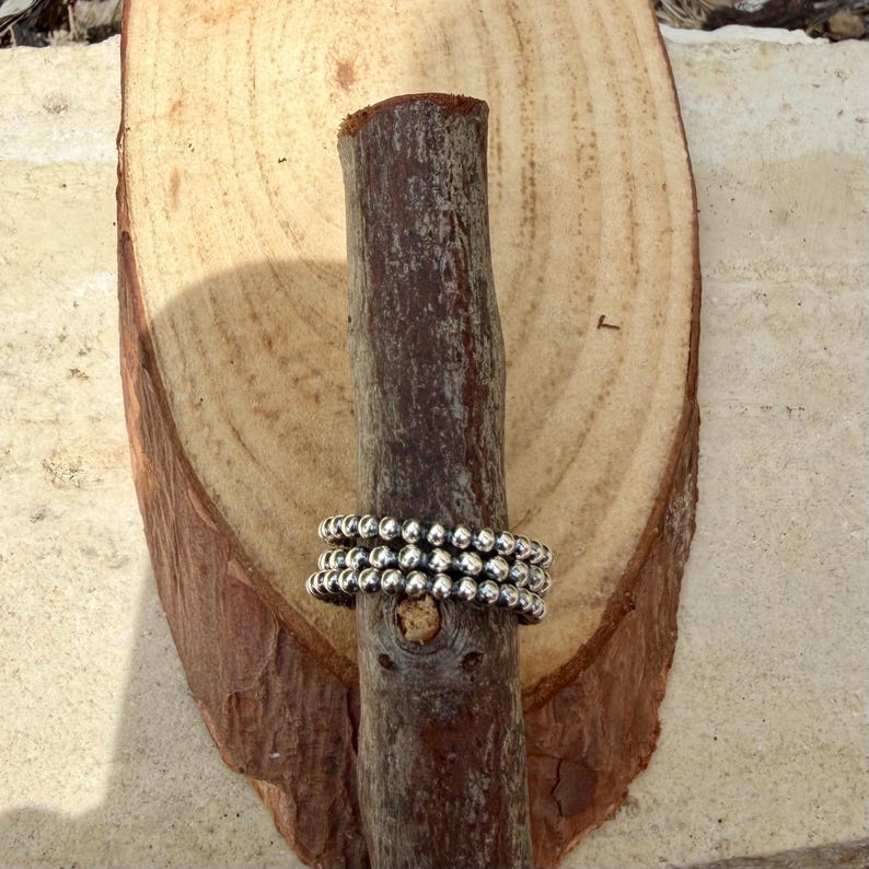 Puede incluir: Tres anillos plateados con cuentas apilados sobre un palo de madera marr&oacute;n oscuro, sobre un fondo de madera marr&oacute;n claro. Los anillos est&aacute;n hechos de peque&ntilde;as cuentas redondas. La madera tiene un aspecto natural y texturizado.