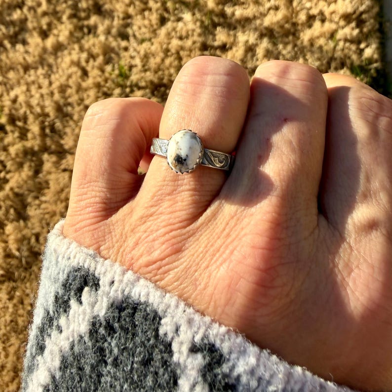 May include: A silver ring with an oval, white gemstone with black and brown inclusions. The ring has a decorative band with etched floral patterns. The ring is worn on a finger, with a patterned gray and white sweater sleeve visible.