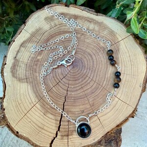 May include: A silver-toned metal chain necklace featuring a single round black stone pendant at the center and a series of five small, round, brown and black tiger eye beads integrated into one side of the chain, secured with a lobster claw clasp.