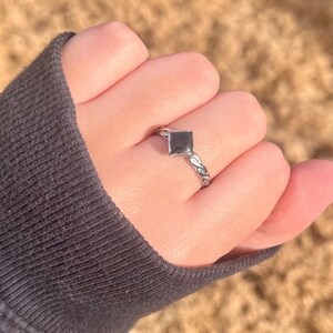 May include: A silver ring with a square, black gemstone. The band features a delicate, swirling design. The ring is shown on a hand, with a dark gray sleeve in the background.