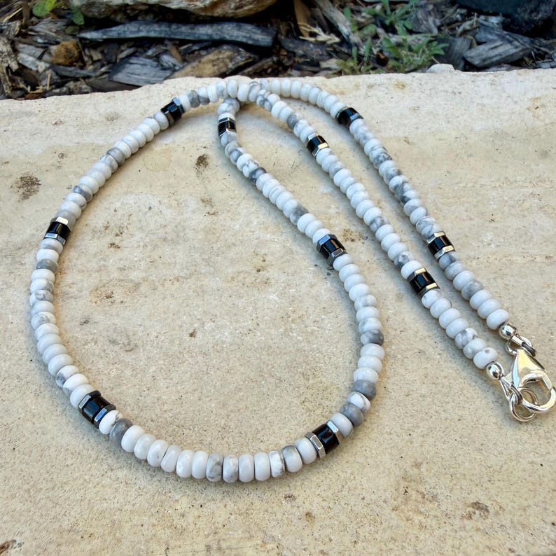 May include: A white and gray beaded necklace with black accents. The necklace features round, marbled beads interspersed with black and silver-toned metal spacers. A silver-colored clasp secures the necklace.