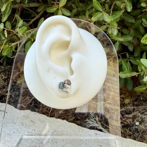 May include: A small, silver-toned heart-shaped stud earring featuring an intricate, engraved floral or scrollwork pattern on its surface. The earring is displayed on a white, ear-shaped mannequin stand made of a smooth, matte material.