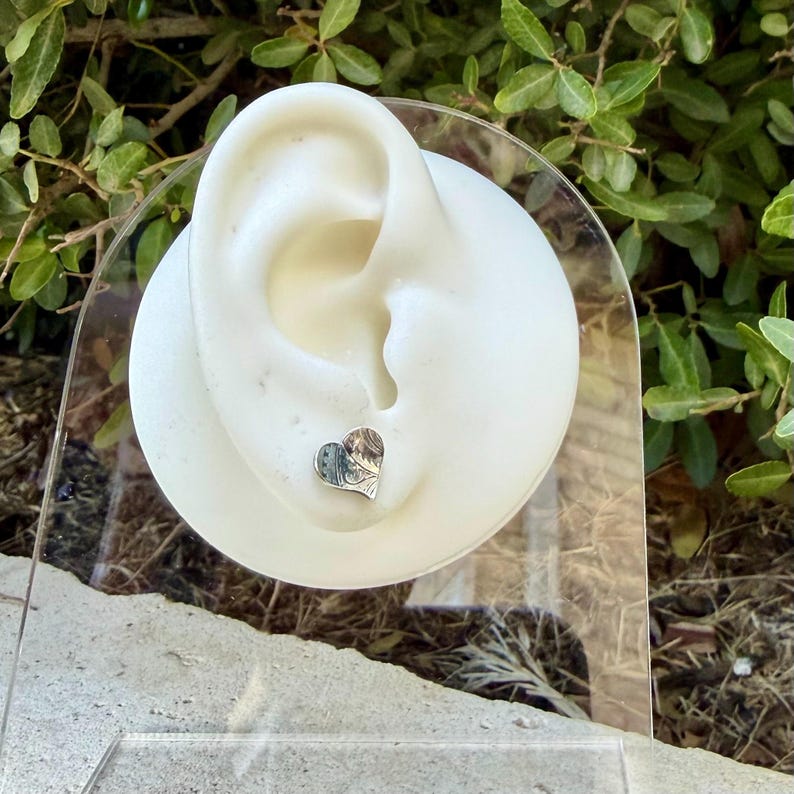 May include: A small, silver-toned heart-shaped stud earring featuring an intricate, engraved floral or scrollwork pattern on its surface. The earring is displayed on a white, ear-shaped mannequin stand made of a smooth, matte material.