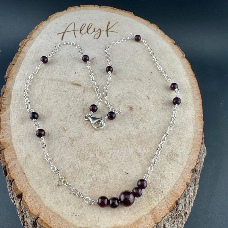 May include: A silver chain necklace with dark red garnet beads. The necklace features a lobster clasp closure and is displayed on a wooden surface. The word "Allyk" is written in cursive on the wood.