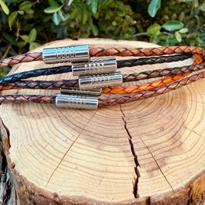 May include: A set of five braided leather bracelets in shades of brown, black, and tan. Each bracelet features a cylindrical, ribbed silver-tone metal clasp. The bracelets are arranged in a stacked, parallel formation, showcasing the texture.