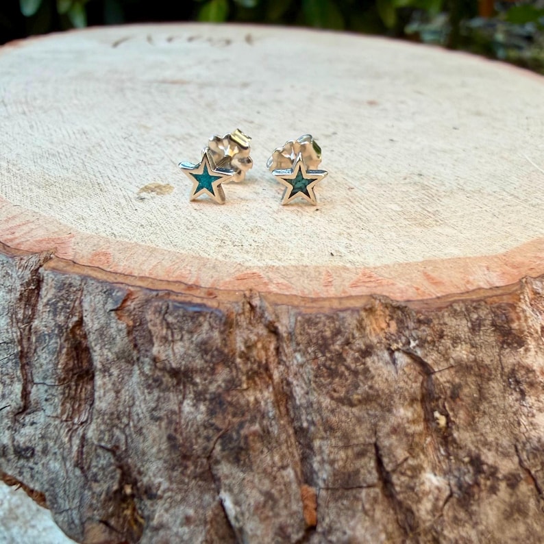May include: A pair of star-shaped stud earrings. Each earring features a silver-colored metal frame with a turquoise-colored center. The earrings are displayed on a natural wood surface.