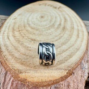 May include: A silver-colored ring with a chain link design. The ring is made of a shiny metal and has a thick band. The chain link pattern is repeated around the entire ring. The ring is displayed on a wooden surface.