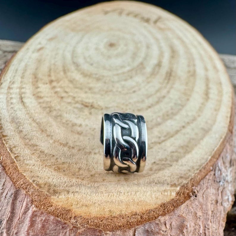 May include: A silver-colored ring with a chain link design. The ring is made of a shiny metal and has a thick band. The chain link pattern is repeated around the entire ring. The ring is displayed on a wooden surface.
