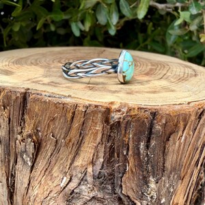 Puede incluir: Un anillo de plata con una banda de metal trenzado y una piedra turquesa lisa de forma ovalada con vetas de matriz de color marr&oacute;n oscuro. La piedra est&aacute; engastada en un sencillo bisel de plata y la banda consta de varios hilos de plata retorcidos.