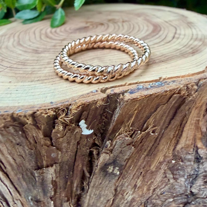 May include: Two gold-colored, twisted rope design rings. The rings are stacked on top of each other. The rings are on a wooden surface with green leaves in the background.