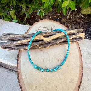 May include: Turquoise beaded necklace with a mix of small square and round beads. The necklace features larger, round turquoise beads interspersed with silver-colored spacers. The necklace is displayed on a wooden surface with the text "Ally K".
