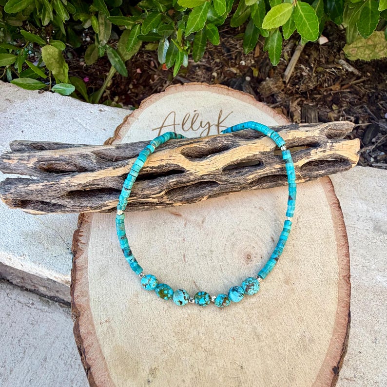 May include: Turquoise beaded necklace with a mix of small square and round beads. The necklace features larger, round turquoise beads interspersed with silver-colored spacers. The necklace is displayed on a wooden surface with the text "Ally K".