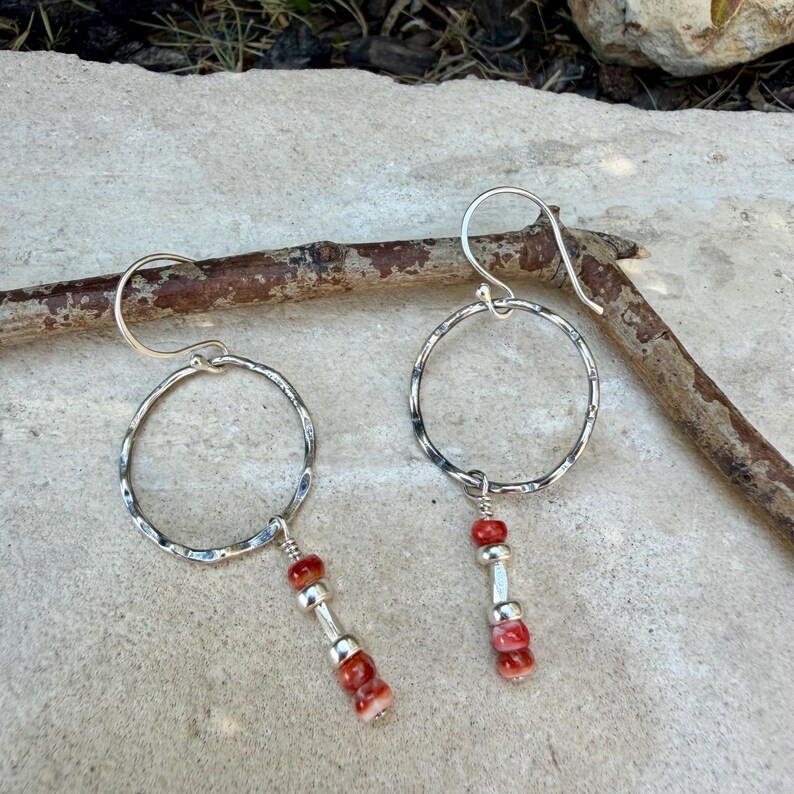 May include: A pair of silver-toned earrings featuring a hammered circular hoop design. Each earring hangs from a curved ear wire and includes a dangling vertical accent composed of two small red beads separated by a central silver-toned spacer bead.