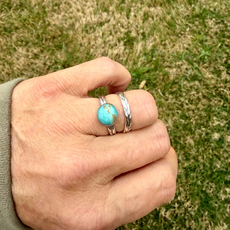May include: A hand wearing two silver rings. One ring features a round turquoise stone, and the other has a textured design. The rings are set against a background of green grass.