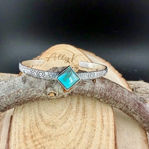 Puede incluir: Una pulsera de plata con una piedra turquesa cuadrada en el centro. La pulsera tiene un patr&oacute;n floral y el texto "AllyK" es visible en la superficie de madera detr&aacute;s de la pulsera.