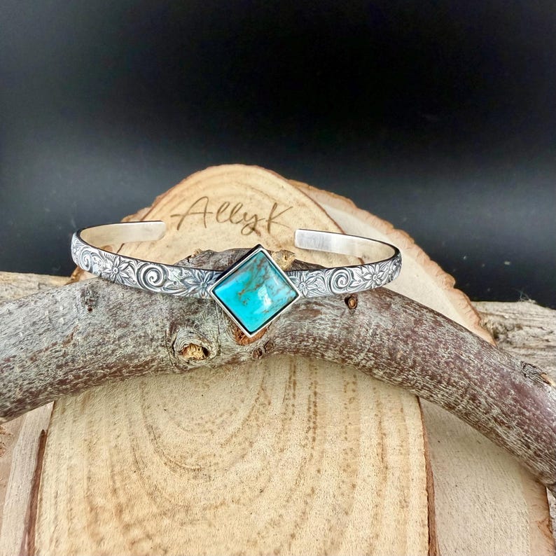 Puede incluir: Una pulsera de plata con una piedra turquesa cuadrada en el centro. La pulsera tiene un patr&oacute;n floral y el texto "AllyK" es visible en la superficie de madera detr&aacute;s de la pulsera.