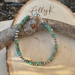 May include: A turquoise and silver beaded bracelet. The bracelet has small, round turquoise beads and silver beads. The clasp is silver, and the bracelet is displayed on a wooden surface. The name "Allyk" is written in cursive.