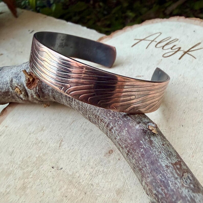 May include: A copper cuff bracelet with a textured, wave-like pattern. The bracelet is displayed on a wooden branch against a light wood background. The bracelet's design features a dark, oxidized interior and a polished exterior.