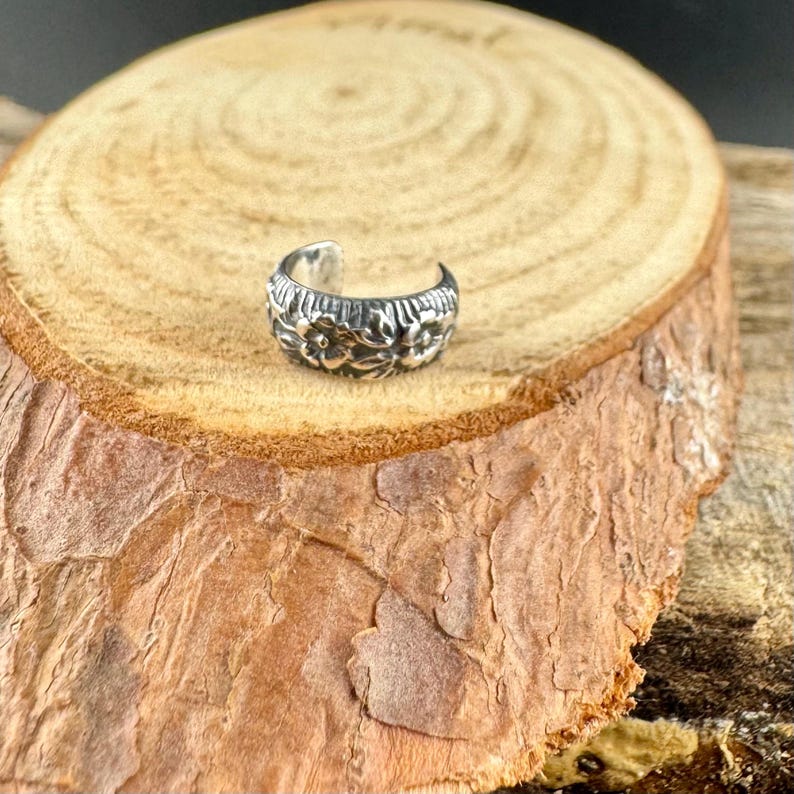 May include: A silver-colored ear cuff with a floral design. The cuff is curved and wraps around the ear. The floral pattern is detailed and raised, adding texture to the piece. The cuff is displayed on a wooden surface.