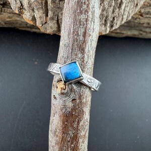 May include: A silver ring with a square, blue gemstone. The band features an etched design. The ring is displayed on a piece of wood, showcasing its details and craftsmanship. A close-up shot highlights the ring's features.