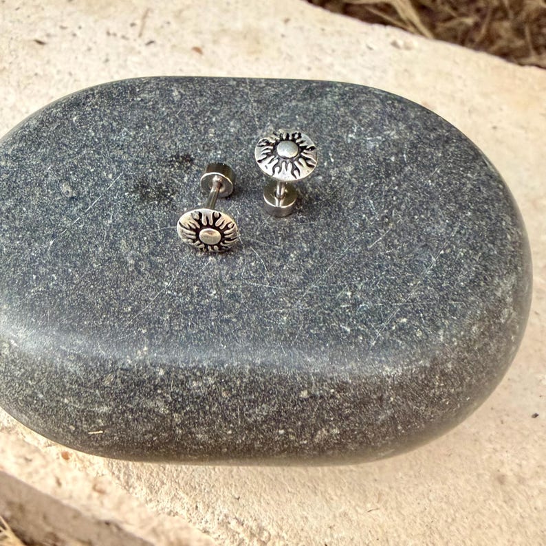 May include: A pair of silver-toned stud earrings featuring a circular design with embossed sun rays radiating from a central dot. The earrings have a metallic finish with darkened recessed areas to highlight the sunburst pattern detail.