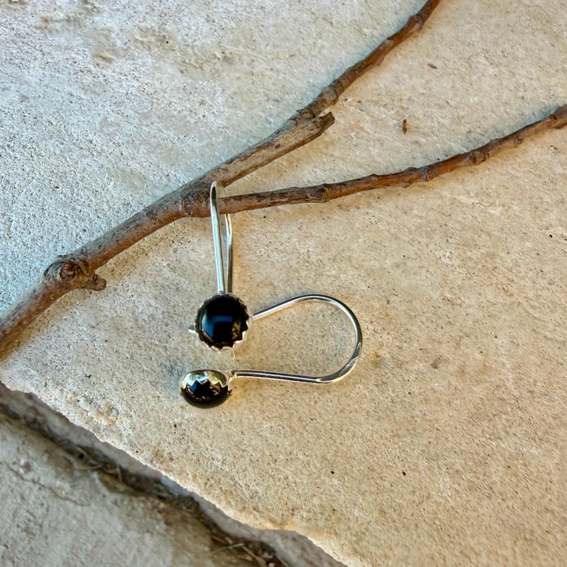 May include: A pair of silver earrings featuring round, faceted black stones set in a scalloped metal bezel. The earrings have a curved, open-loop wire design that hooks through the ear, showcasing a polished metallic finish and dark gems.