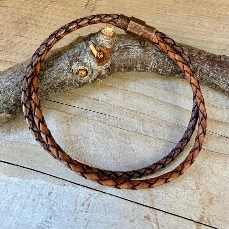 Puede incluir: Una pulsera de cord&oacute;n de cuero trenzado marr&oacute;n con un cierre cil&iacute;ndrico de metal en tono cobre. La pulsera tiene forma circular y muestra una textura tejida de m&uacute;ltiples hilos a lo largo de todo el cord&oacute;n, terminada con un conector met&aacute;lico liso.