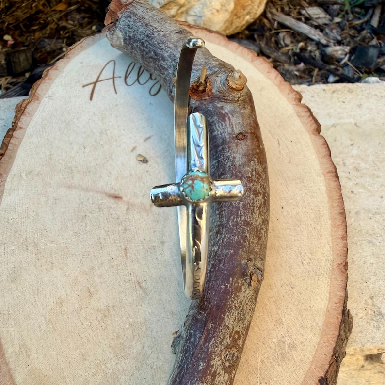 May include: A silver cuff bracelet with a cross design and a turquoise stone center. The bracelet is displayed on a wooden log. The word "Always" is written on the wooden surface.