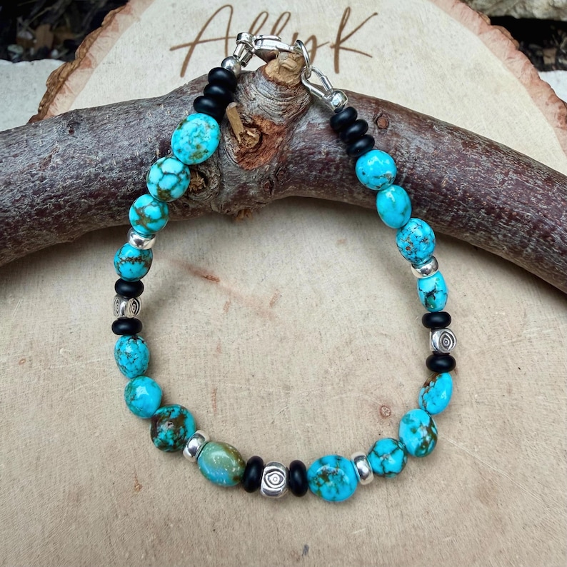 May include: Turquoise beaded bracelet with silver accents and black beads. The bracelet features round, turquoise-colored beads interspersed with silver-colored spacers and black beads. The clasp is silver-colored. The bracelet is displayed on a wooden surface.