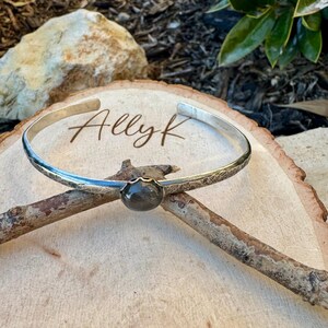 May include: A silver cuff bracelet with a dark gemstone center. The bracelet has a textured band and the text "Ally K" is visible on the wooden surface. The bracelet is displayed on a wooden surface.