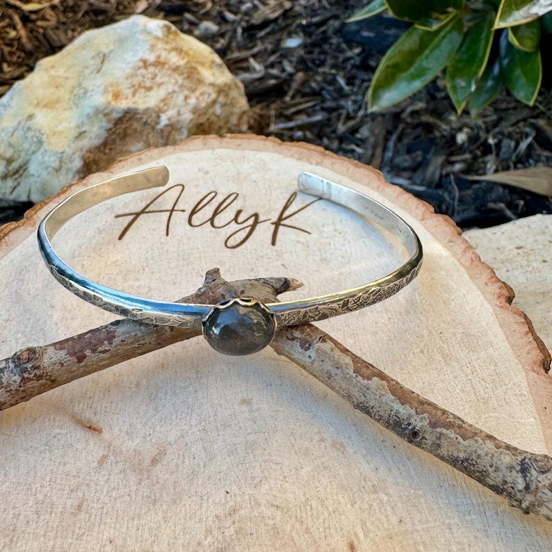 May include: A silver cuff bracelet with a dark gemstone center. The bracelet has a textured band and the text "Ally K" is visible on the wooden surface. The bracelet is displayed on a wooden surface.