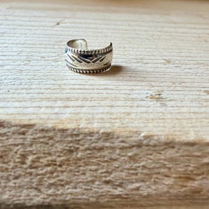 May include: A silver-colored metal ear cuff featuring a wide band design with an open back. The surface of the ear cuff is decorated with a series of etched geometric patterns, including horizontal lines and small triangular shapes.