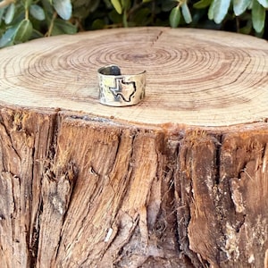 May include: A silver-toned metal ring featuring an open band design with a cutout silhouette of the state of Texas. The surface of the ring has a hammered, textured finish with darkened detailing around the edges of the state shape.
