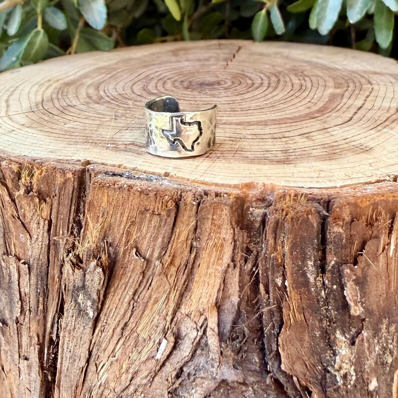 May include: A silver-toned metal ring featuring an open band design with a cutout silhouette of the state of Texas. The surface of the ring has a hammered, textured finish with darkened detailing around the edges of the state shape.