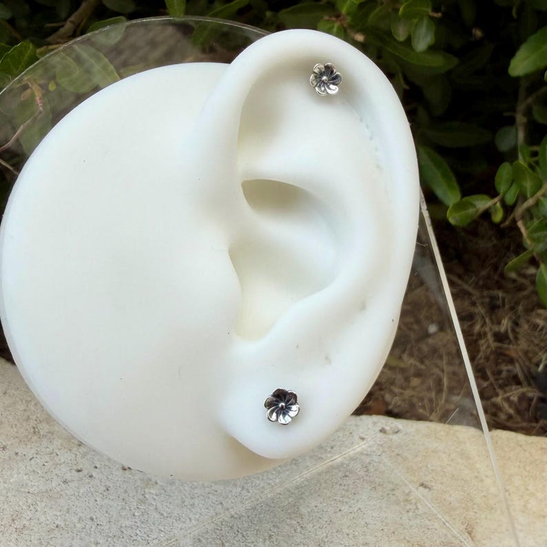 May include: Two small, silver-toned flower-shaped stud earrings are displayed on a white ear-shaped mannequin. Each earring features a detailed, five-petaled floral design with a central dot, positioned in the upper and lower ear lobes.