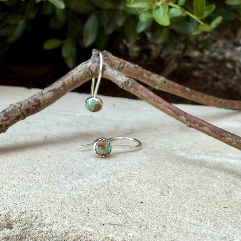 May include: A pair of silver drop earrings featuring round, turquoise-colored stones with brown matrix patterns. One earring hangs from a thin wooden branch, while the other rests on a light-colored stone surface, showcasing the metal settings.