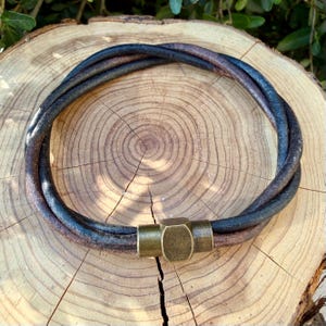 May include: A multi-strand bracelet composed of dark brown and black leather cords twisted together in a circular shape, secured by a cylindrical antique brass magnetic clasp with a hexagonal centre detail and a smooth metallic finish.
