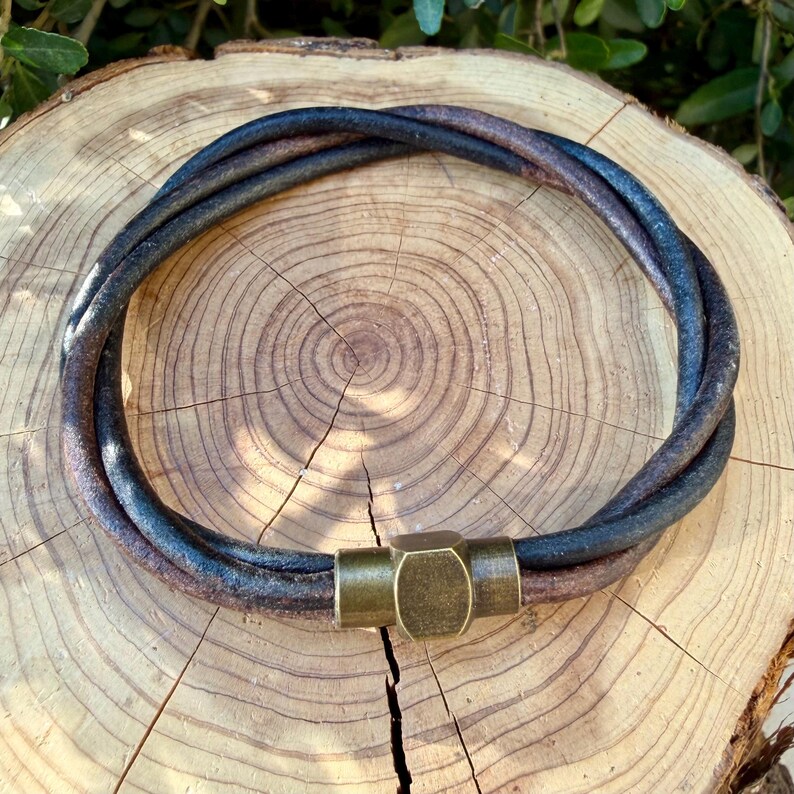 May include: A multi-strand bracelet composed of dark brown and black leather cords twisted together in a circular shape, secured by a cylindrical antique brass magnetic clasp with a hexagonal centre detail and a smooth metallic finish.