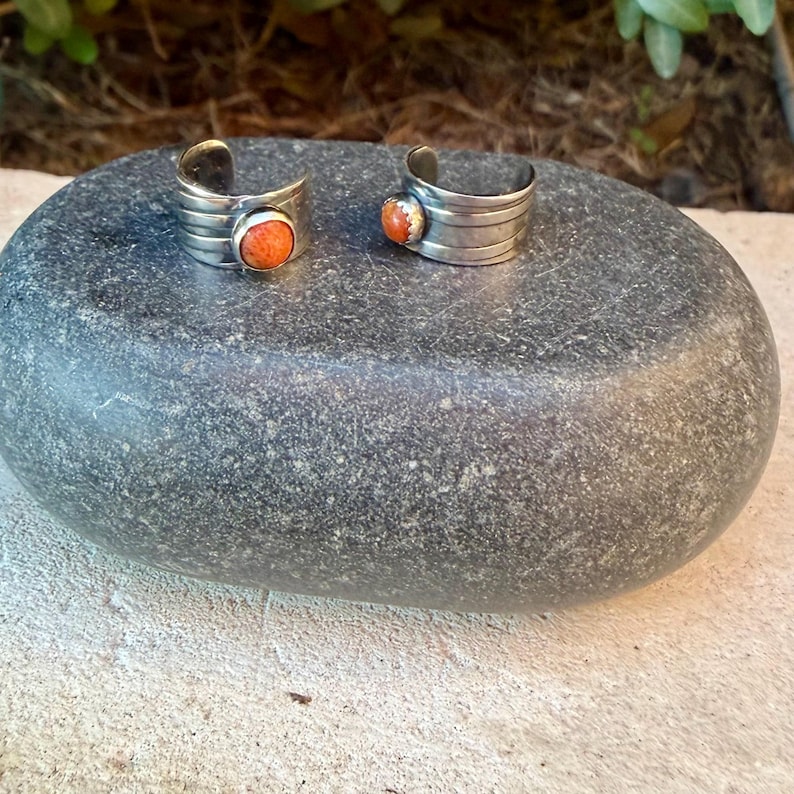 May include: Two silver-toned open-ended rings, each featuring a single round, textured orange stone set in a smooth bezel. The bands are constructed with multiple horizontal ridges that wrap around the finger, creating a layered appearance.