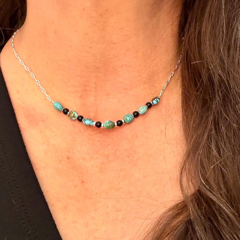 May include: A delicate necklace featuring turquoise and black beads strung on a silver chain. The turquoise beads are a vibrant blue-green color, and the black beads are interspersed throughout the design. The necklace is a simple yet elegant piece of jewelry.