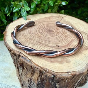 May include: A twisted metal cuff bracelet with copper and silver tones. The bracelet is made of two strands of metal that are intertwined. The bracelet is displayed on a wooden surface with visible wood grain.