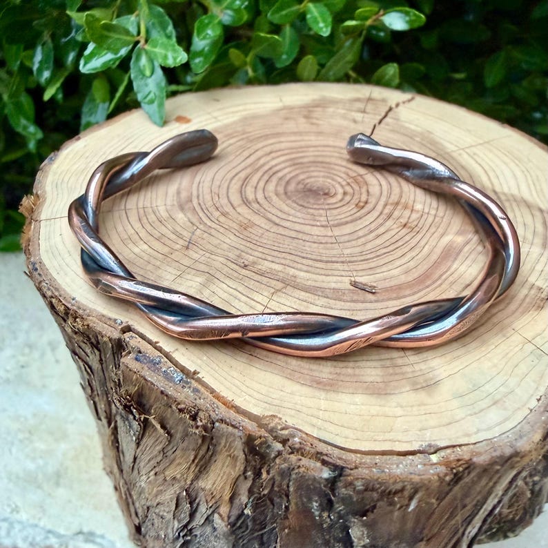 May include: A twisted metal cuff bracelet with copper and silver tones. The bracelet is made of two strands of metal that are intertwined. The bracelet is displayed on a wooden surface with visible wood grain.