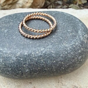 May include: Two gold-colored, twisted rope rings rest on a gray stone. The rings are made of a shiny metal and have a textured, braided appearance. The rings are likely for the fingers and are a jewelry item.