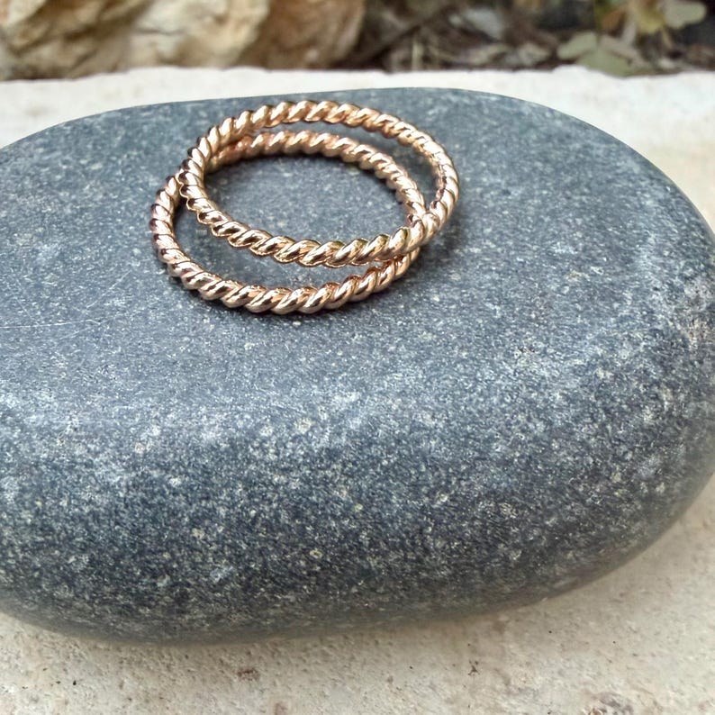May include: Two gold-colored, twisted rope rings rest on a gray stone. The rings are made of a shiny metal and have a textured, braided appearance. The rings are likely for the fingers and are a jewelry item.