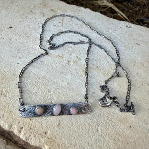 May include: A silver-toned metal necklace featuring a rectangular bar pendant with an etched swirl pattern. The pendant is set with three smooth, oval-shaped, pale pink stones. The necklace includes a delicate chain with a lobster claw clasp.