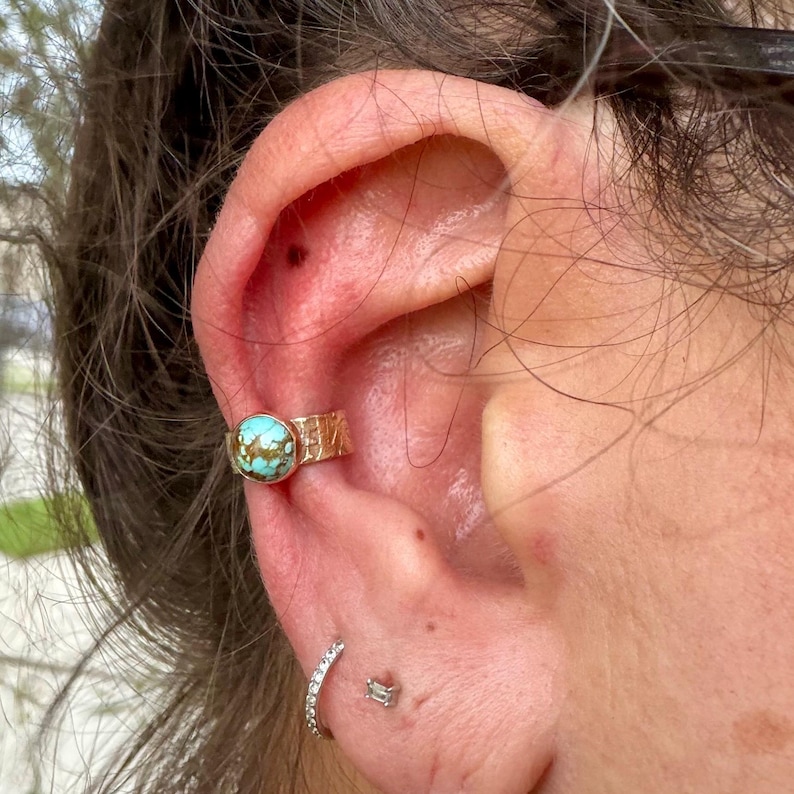 May include: A gold-colored ear cuff with a turquoise stone and textured design. The cuff is worn on the upper ear. A small, silver hoop earring and a small, silver stud earring are also visible.