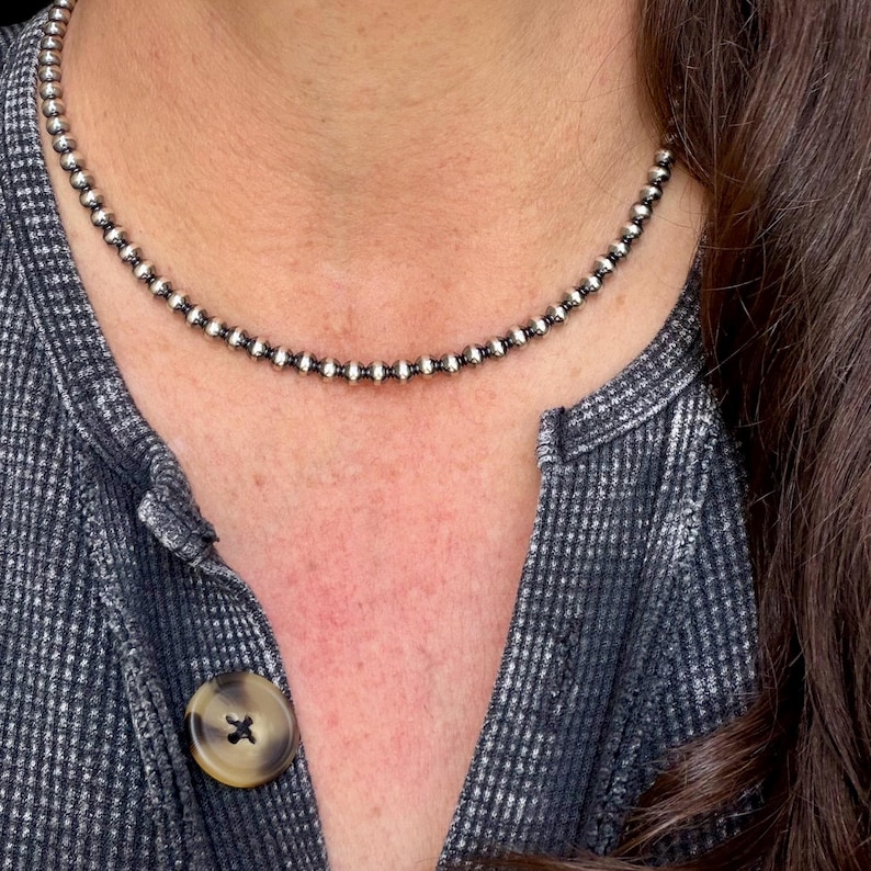 Pu&ograve; includere: Una collana di perline d'argento con piccole perline rotonde. La collana &egrave; indossata su una camicia strutturata grigia e nera con un grande bottone rotondo. La collana &egrave; un gioiello semplice ed elegante.