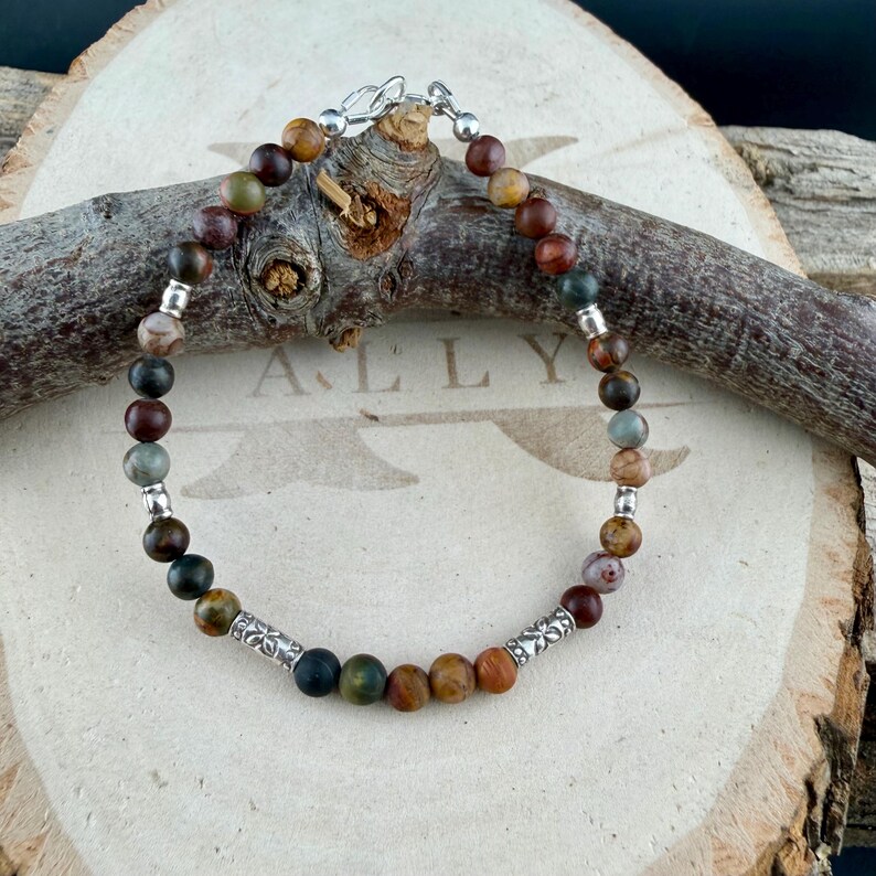 May include: A beaded bracelet featuring round, multi-colored earth-tone stone beads in shades of brown, green, and grey. The bracelet is accented with small silver-toned metal spacers and two cylindrical silver-toned beads with floral engravings.