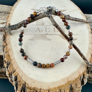 May include: A beaded bracelet featuring round matte earth-toned stones in shades of brown, tan, and muted green, interspersed with small silver-colored metal spacer beads and two ornate silver-colored cylindrical accent beads.